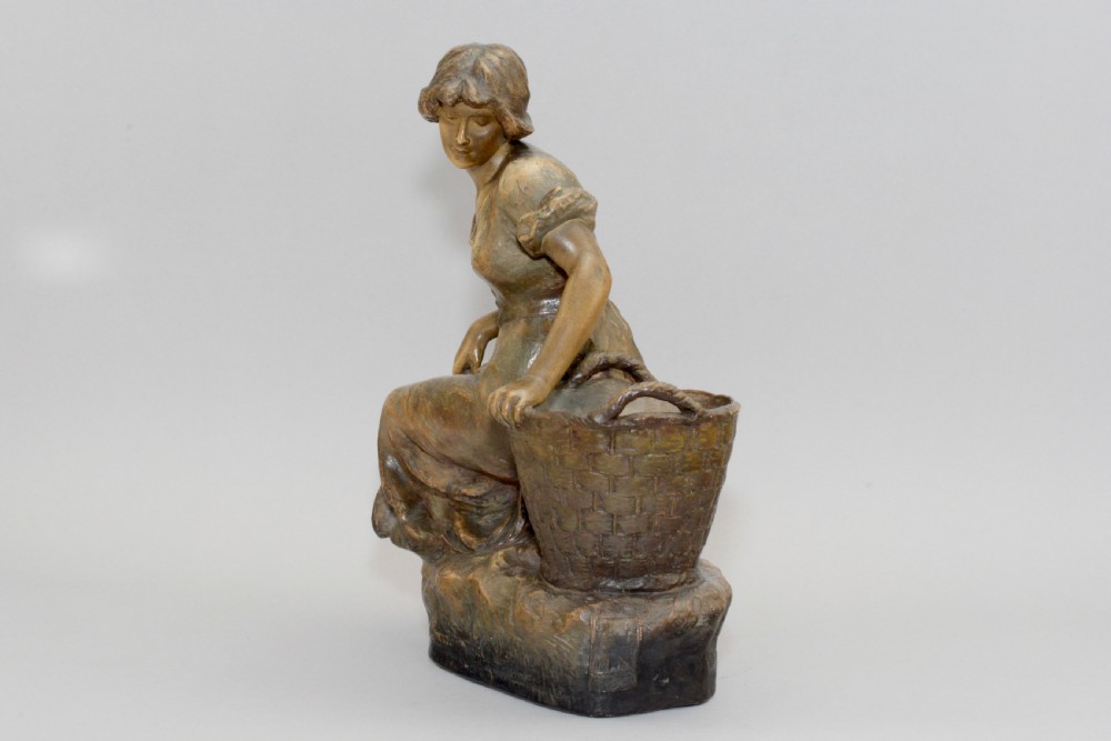 An Art Nouveau terracotta sculpture depicting a seated country maiden in draped clothing beside a woven basket. View IV