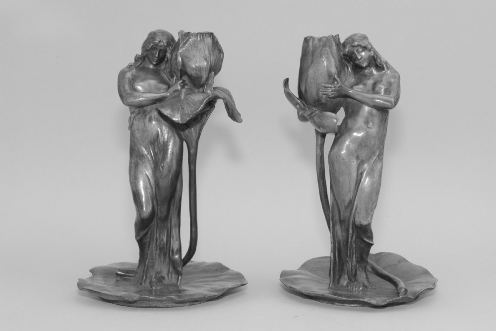 A pair of Art Nouveau pewter candlesticks modelled as draped female figures embracing large flower blossoms on lily pad bases.