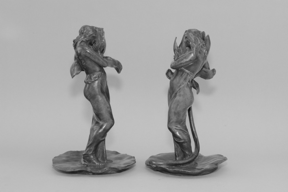A pair of Art Nouveau pewter candlesticks modelled as draped female figures embracing large flower blossoms on lily pad bases. View II