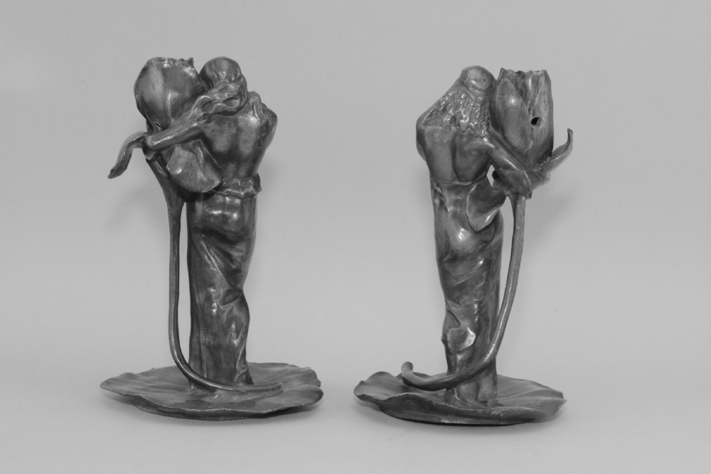 A pair of Art Nouveau pewter candlesticks modelled as draped female figures embracing large flower blossoms on lily pad bases. View III