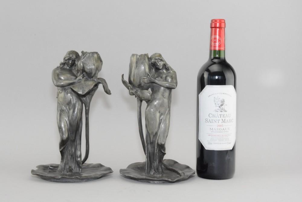 A pair of Art Nouveau pewter candlesticks modelled as draped female figures embracing large flower blossoms on lily pad bases. View VIII