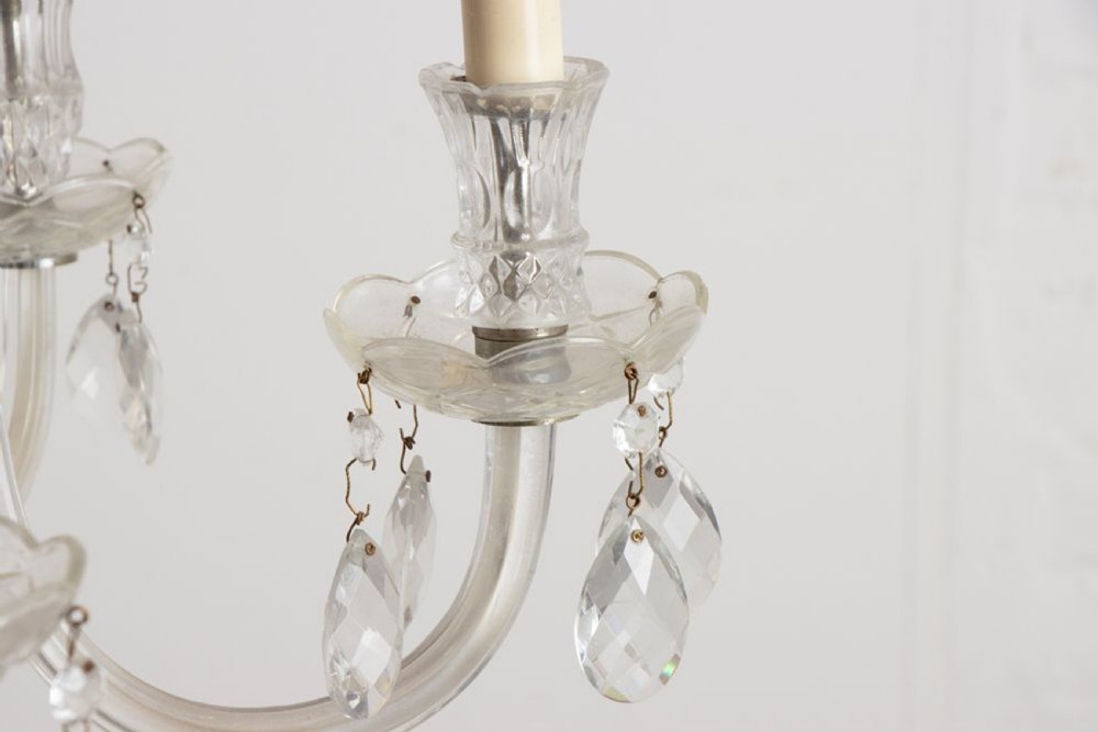 A 1920s clear glass chandelier featuring curved swan-style arms, candle-shaped light sockets, hanging crystal droplets, and a silver ceiling chain. View VI