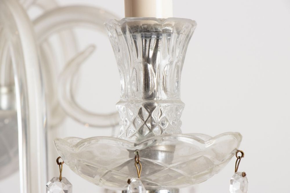 A 1920s clear glass chandelier featuring curved swan-style arms, candle-shaped light sockets, hanging crystal droplets, and a silver ceiling chain. View X