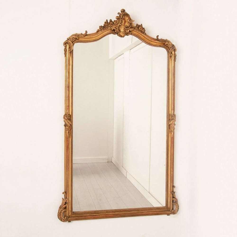 An upright rectangular French giltwood wall mirror featuring a crested top with acanthus leaf ornamentation and subtle red bole showing beneath the gold leaf finish.