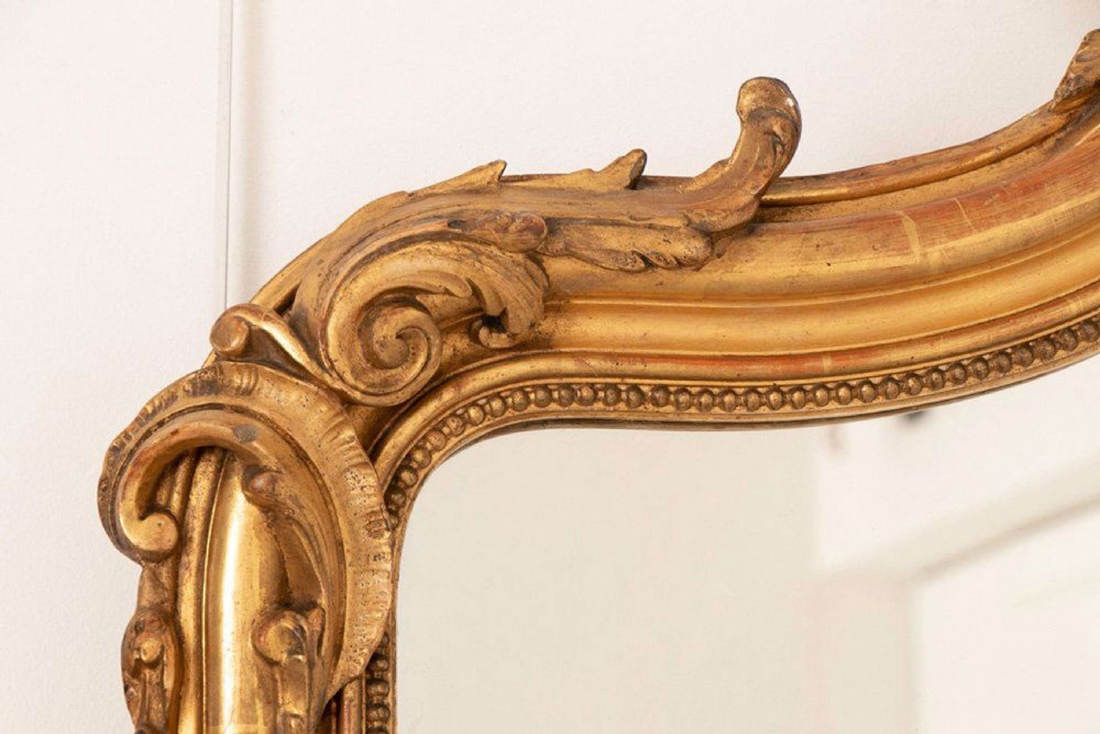 An upright rectangular French giltwood wall mirror featuring a crested top with acanthus leaf ornamentation and subtle red bole showing beneath the gold leaf finish. View IV