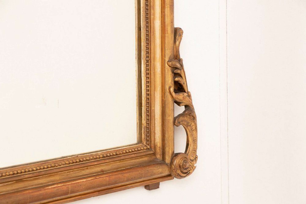 An upright rectangular French giltwood wall mirror featuring a crested top with acanthus leaf ornamentation and subtle red bole showing beneath the gold leaf finish. View V