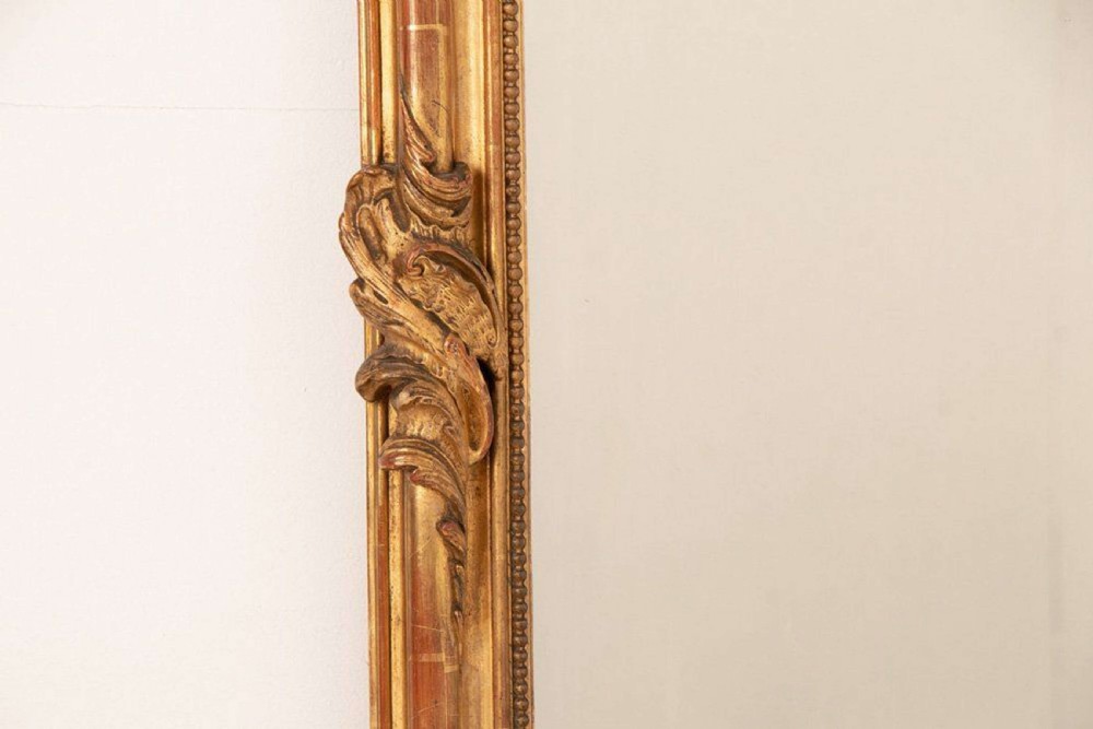 An upright rectangular French giltwood wall mirror featuring a crested top with acanthus leaf ornamentation and subtle red bole showing beneath the gold leaf finish. View VI