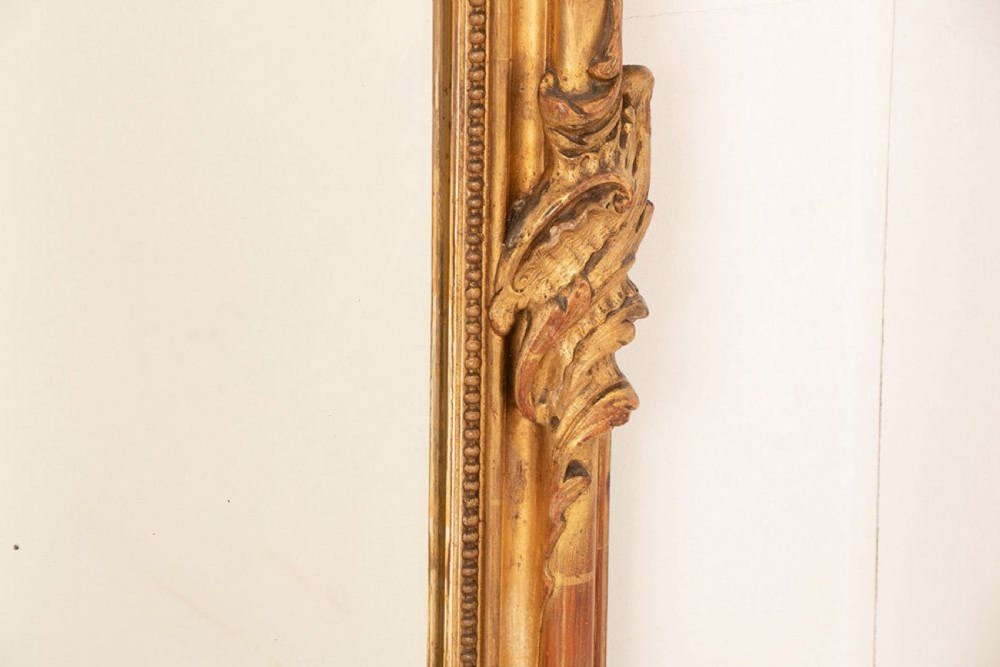 An upright rectangular French giltwood wall mirror featuring a crested top with acanthus leaf ornamentation and subtle red bole showing beneath the gold leaf finish. View VII