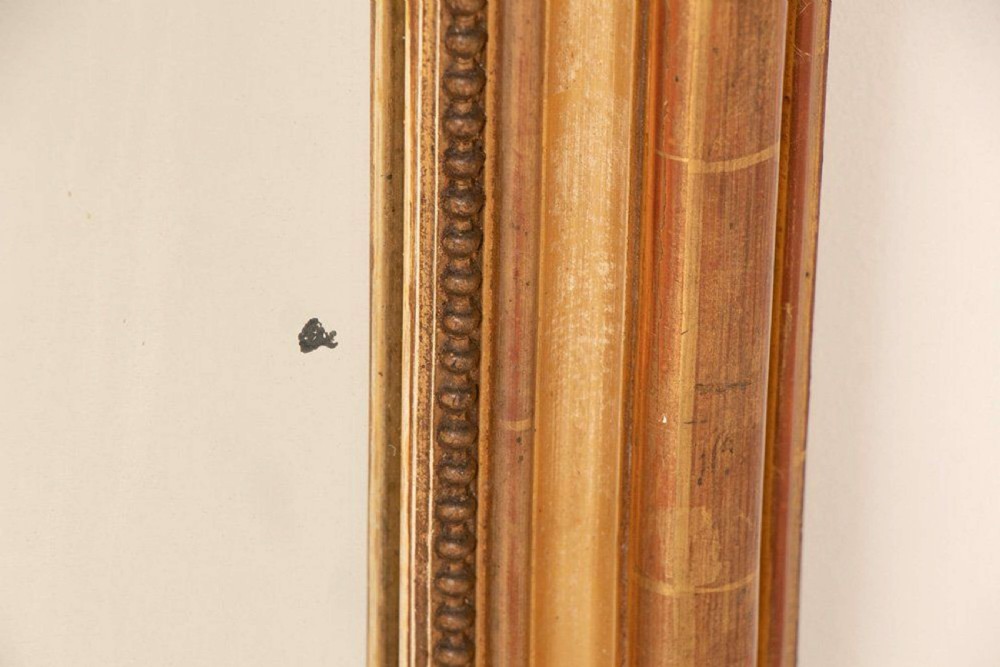 An upright rectangular French giltwood wall mirror featuring a crested top with acanthus leaf ornamentation and subtle red bole showing beneath the gold leaf finish. View VIII