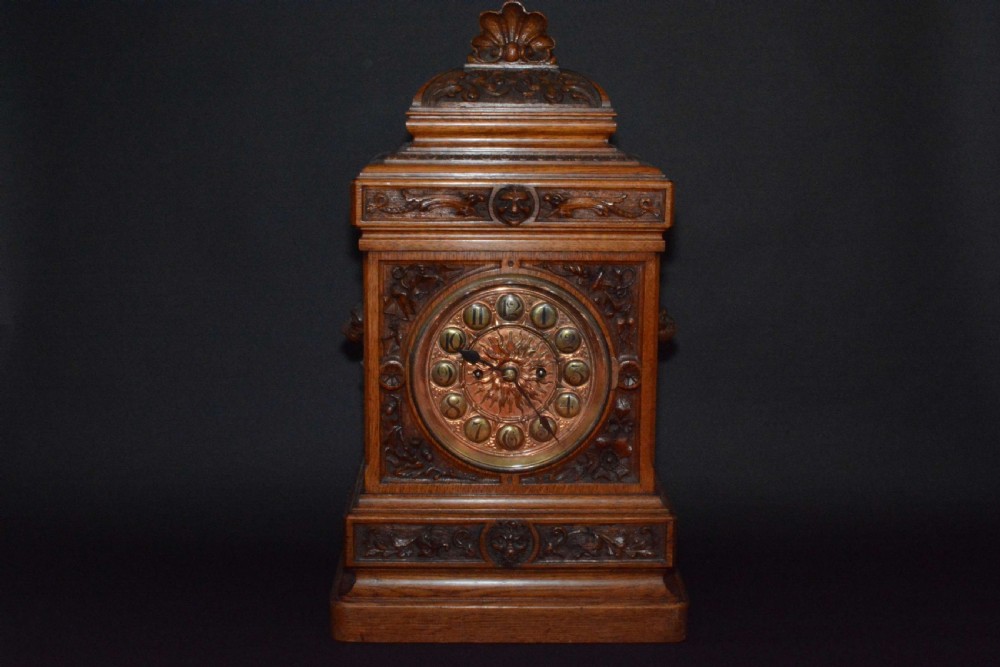 A hand-carved oak mantel clock featuring a central copper dial with raised numerals, flanked by intricate foliate carvings and topped with a shell finial. View III