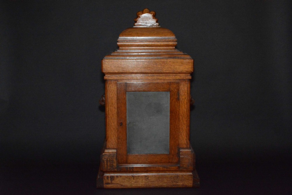 A hand-carved oak mantel clock featuring a central copper dial with raised numerals, flanked by intricate foliate carvings and topped with a shell finial. View V