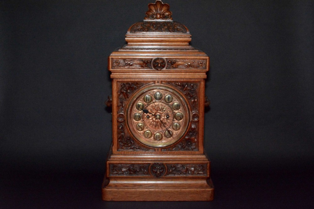 A hand-carved oak mantel clock featuring a central copper dial with raised numerals, flanked by intricate foliate carvings and topped with a shell finial. View VII