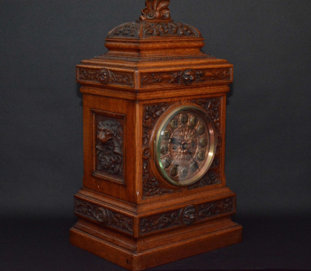 A hand-carved oak mantel clock featuring a central copper dial with raised numerals, flanked by intricate foliate carvings and topped with a shell finial. View X