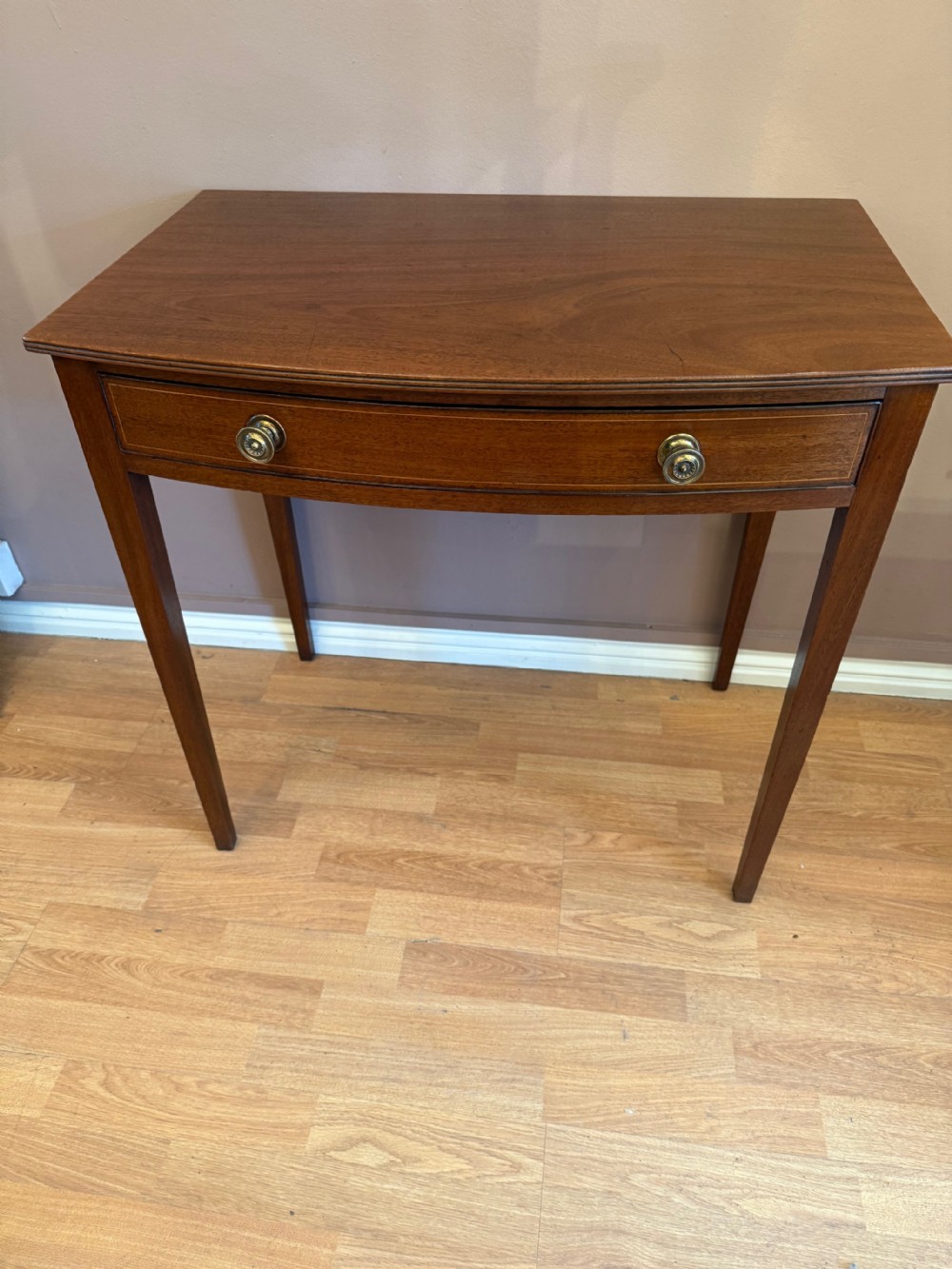 A mahogany bow-fronted side table featuring a single drawer with two circular brass knobs, supported by four tapered square legs.