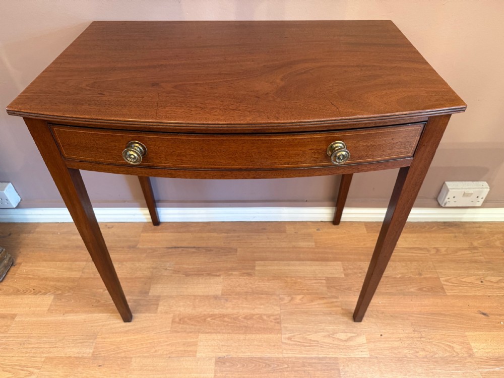 A mahogany bow-fronted side table featuring a single drawer with two circular brass knobs, supported by four tapered square legs. View III