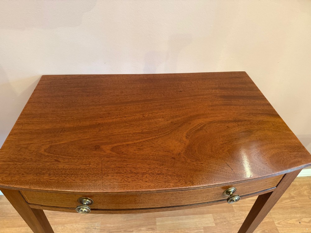 A mahogany bow-fronted side table featuring a single drawer with two circular brass knobs, supported by four tapered square legs. View IV
