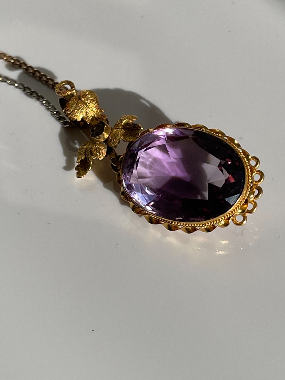 An oval purple faceted amethyst pendant with an ornate foliate gold bail on a delicate chain. View III