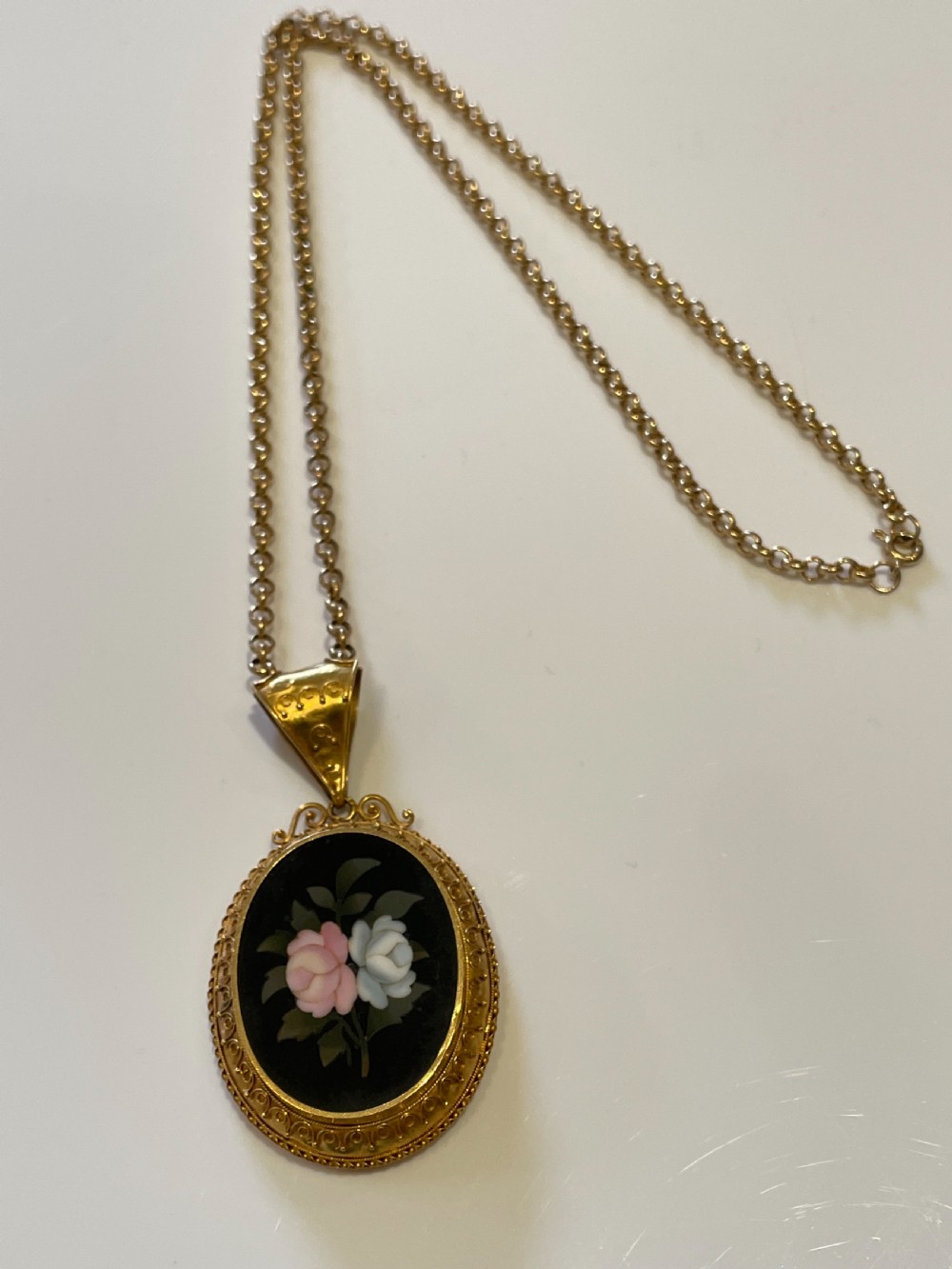An oval 18ct yellow gold pendant featuring a black Pietra Dura hardstone inlay of pink and white roses suspended from a gold chain.