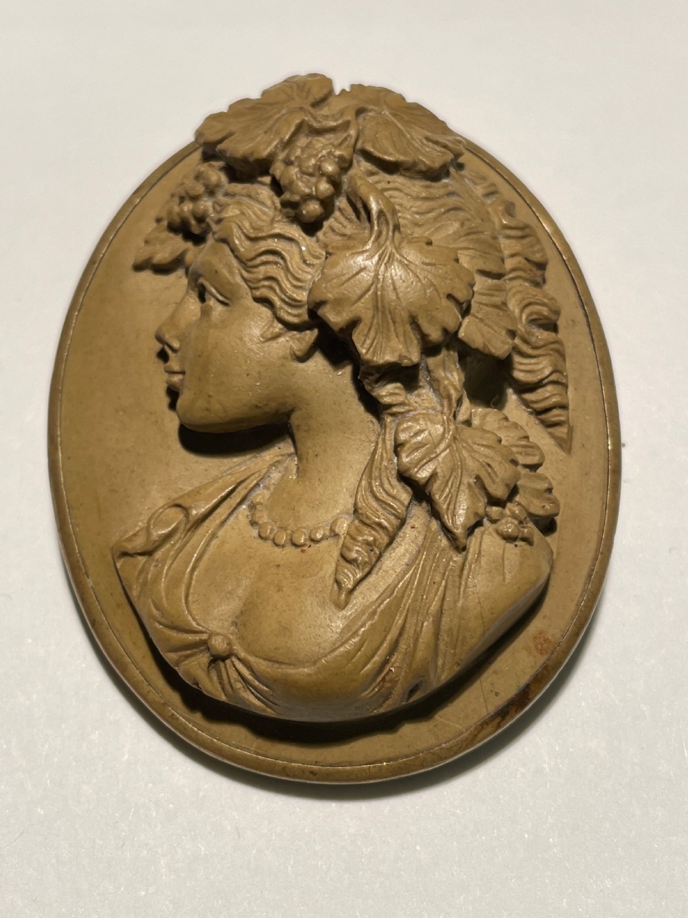 A tan carved lava stone cameo brooch depicting the profile of a classical figure wearing a grapevine wreath.