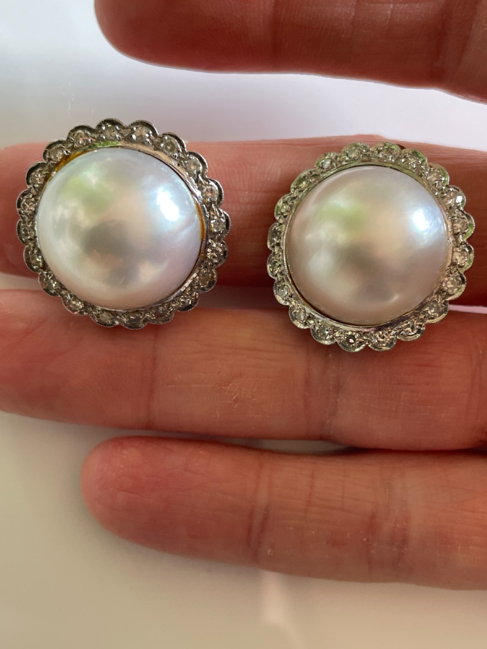 A pair of large circular mid-century earrings featuring central round pearls encircled by scalloped metal halos set with small diamonds.