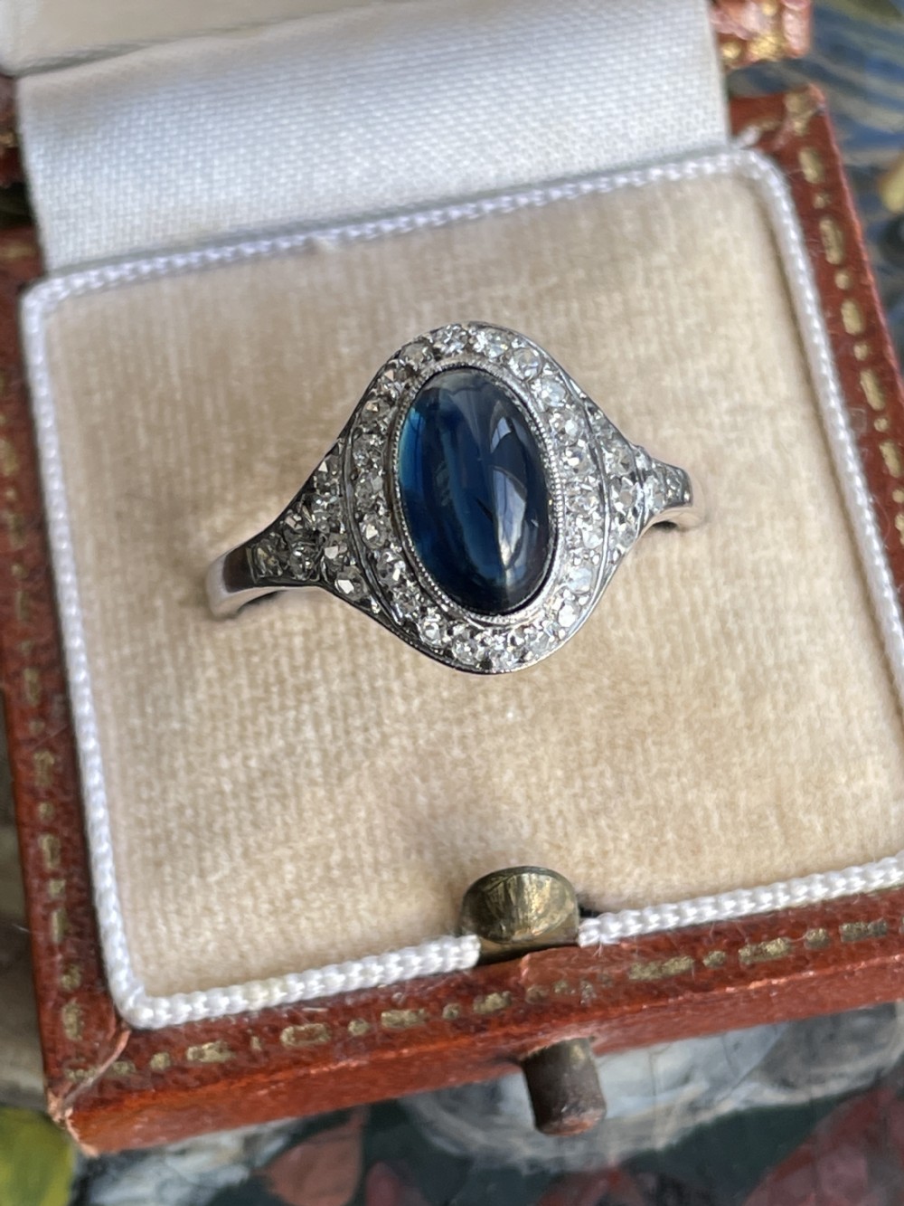 An Art Deco ring featuring an oval cabochon cut deep blue sapphire set within a halo of small diamonds. View IV