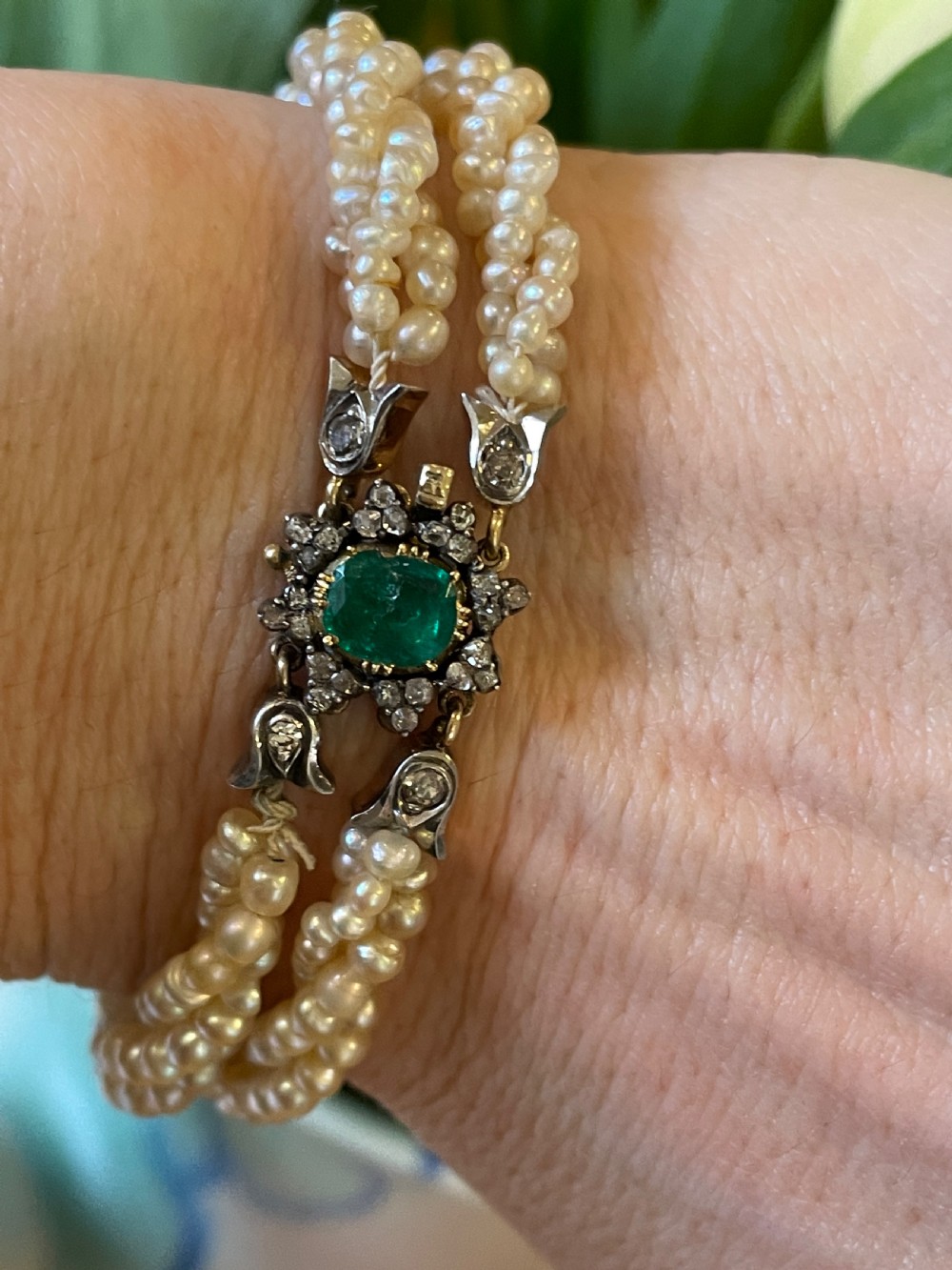 A multi-strand natural white pearl bracelet featuring an oval green emerald centre surrounded by a cluster of diamonds with leaf-shaped caps. View VI