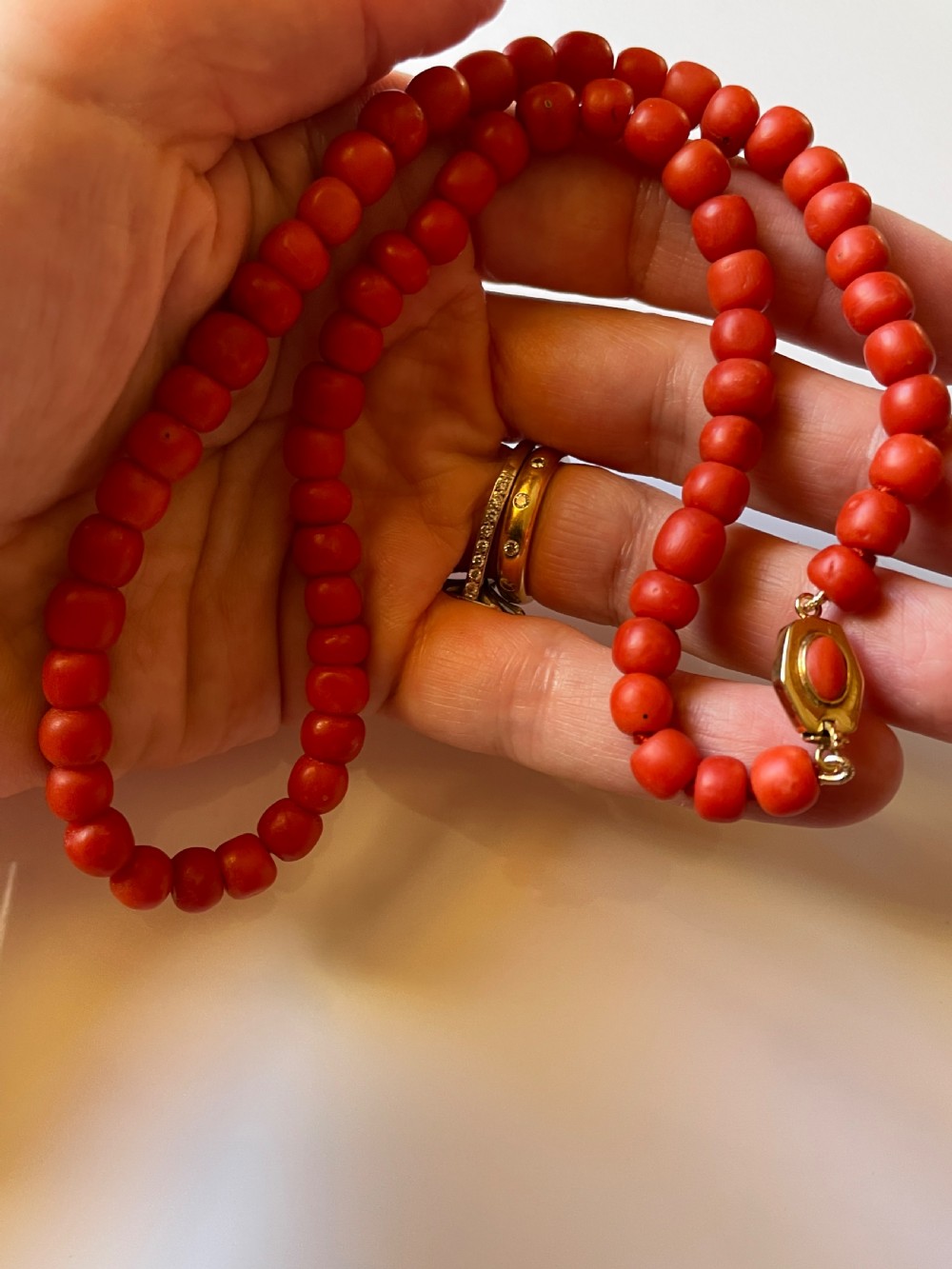 A hand holding a single strand of natural un-dyed red coral beads featuring a gold push clasp.
