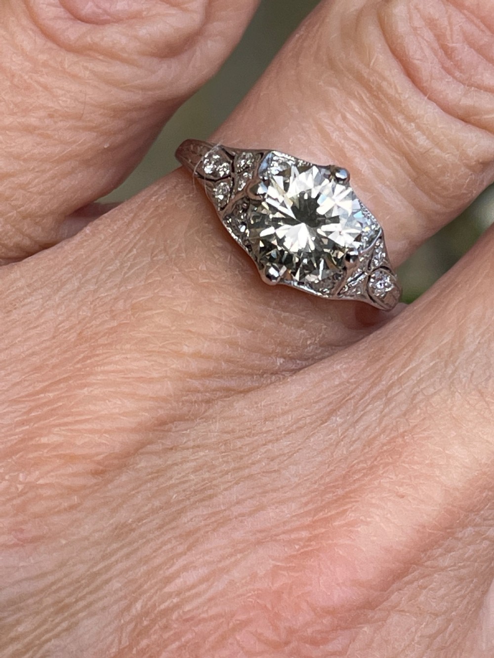 An Art Deco platinum ring featuring a large central round diamond with smaller accent diamonds and leaf motifs engraved into the shoulders. View II