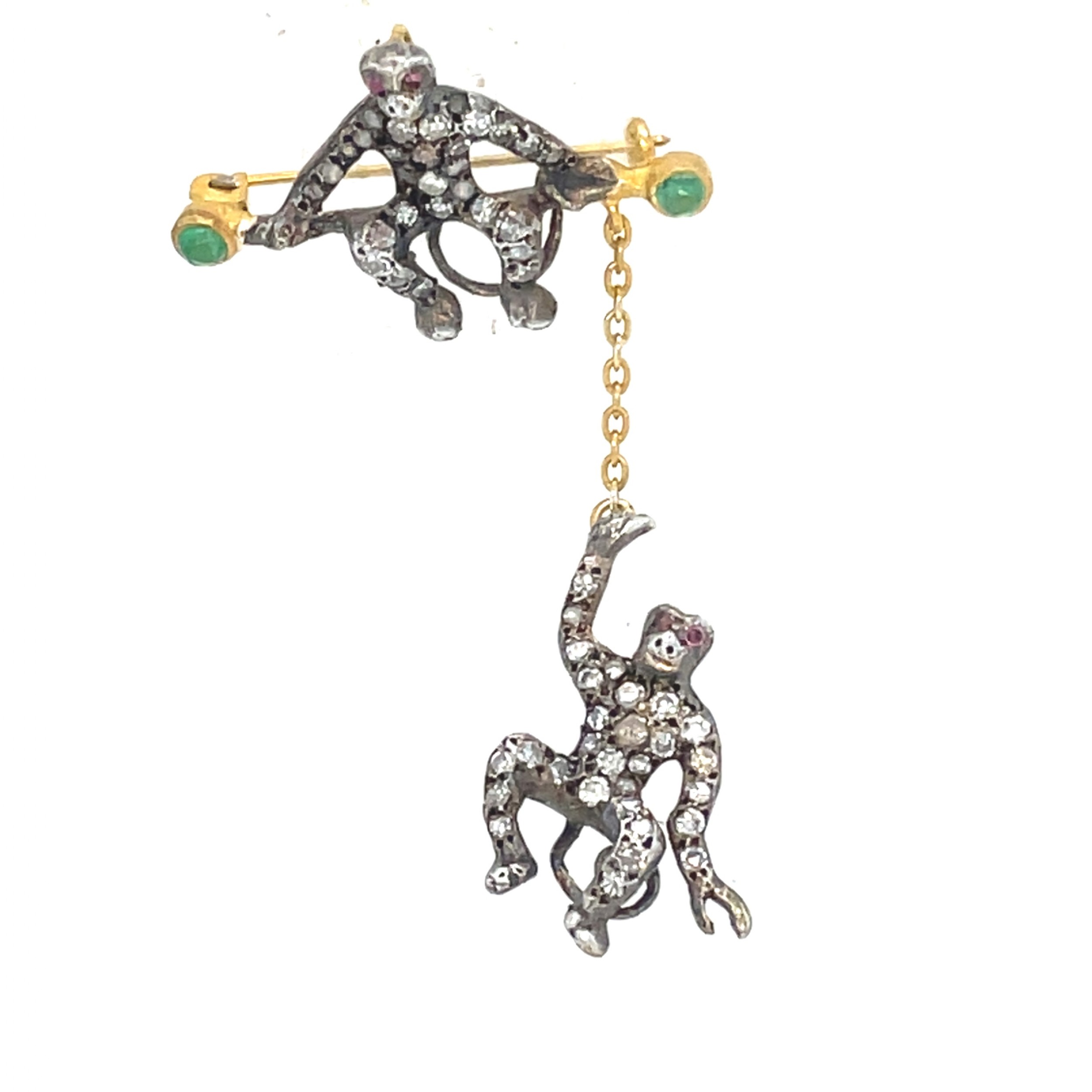 Victorian bar brooch featuring two pave diamond-set monkeys, one perched on a gold bar with green emerald terminals and one dangling below from a chain.