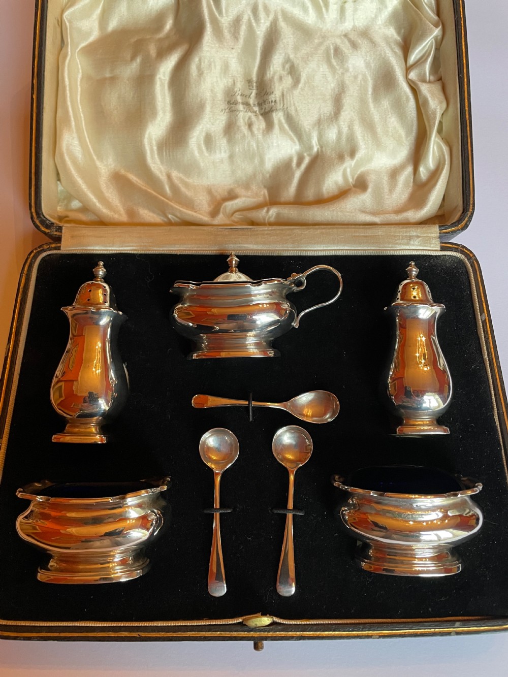 A cased sterling silver condiment set featuring two baluster pepper castors, two open salt cellars, a mustard pot, and three spoons inside a silk-lined box.