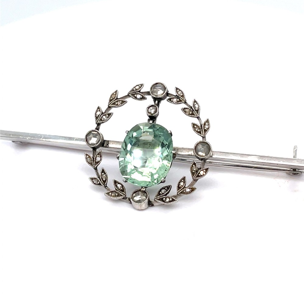 An Edwardian bar brooch featuring a central oval pale green aquamarine encircled by a diamond-set leaf wreath motif.