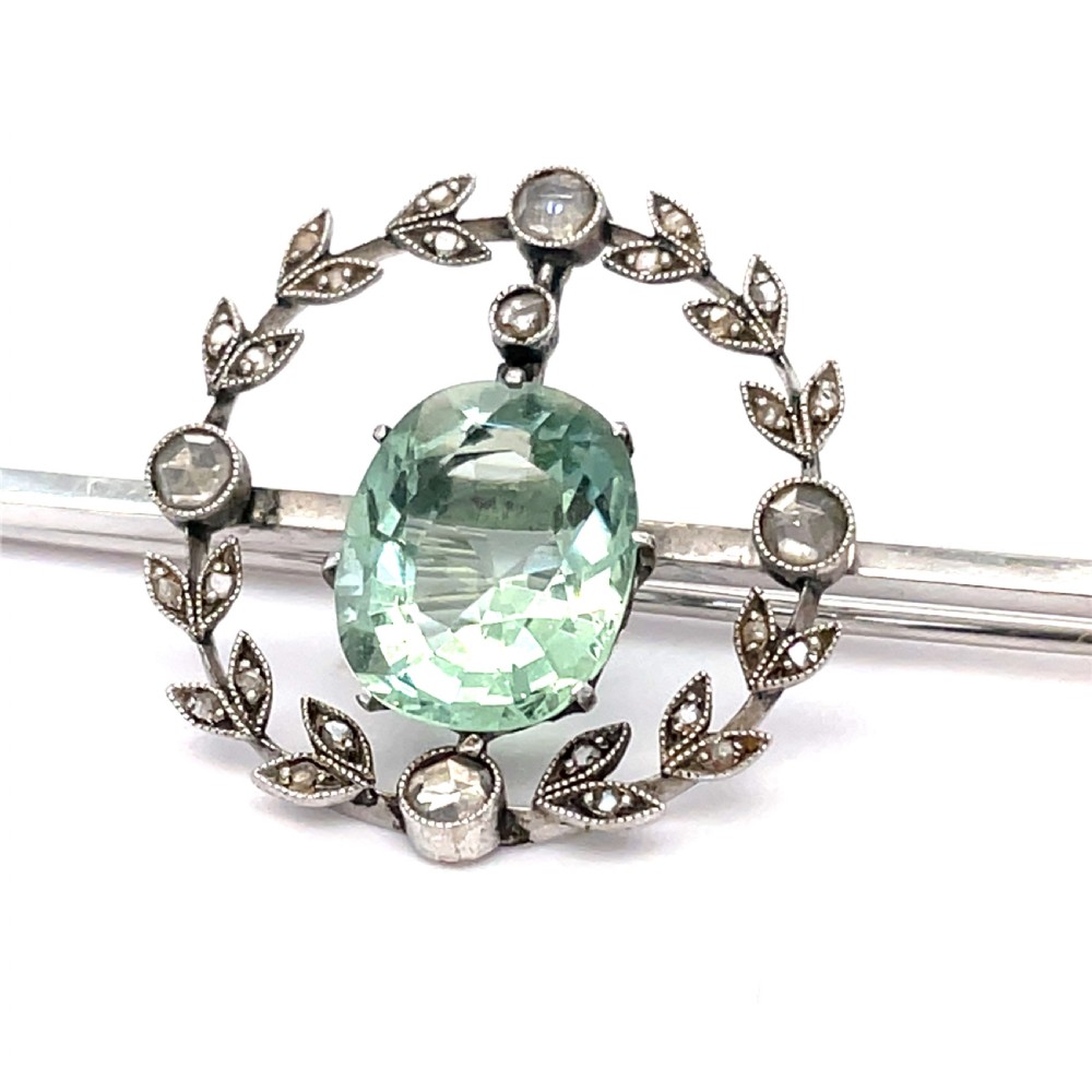 An Edwardian bar brooch featuring a central oval pale green aquamarine encircled by a diamond-set leaf wreath motif. View V