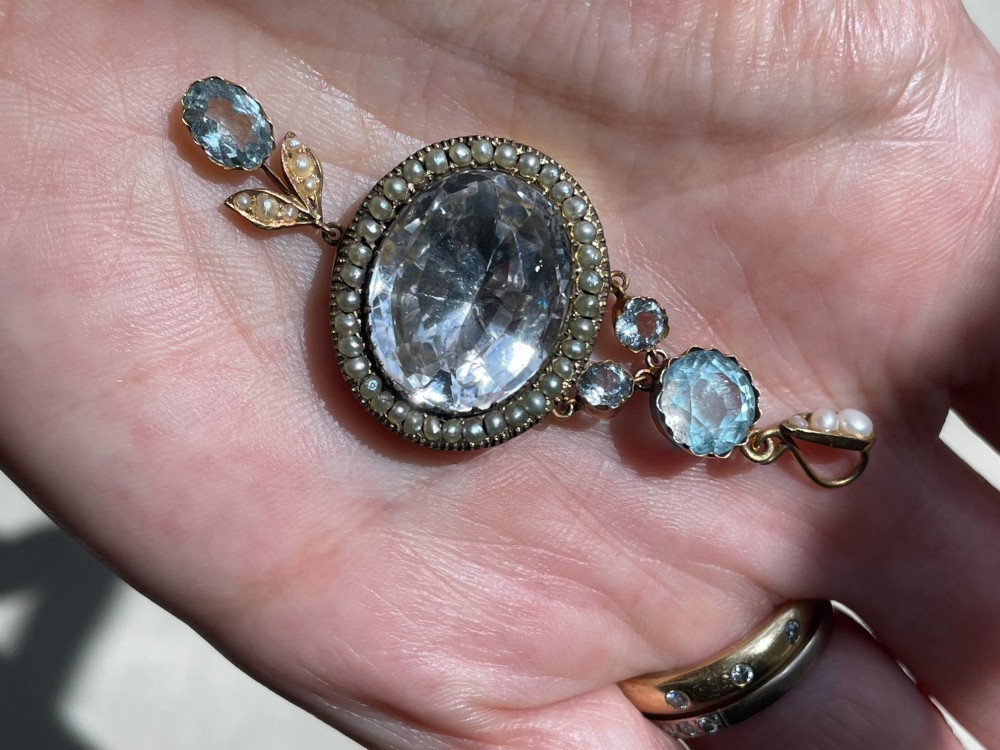 A Victorian gold pendant featuring a large oval rock crystal bordered by split seed pearls, flanked by pale blue aquamarine stones and leaf motifs. View II