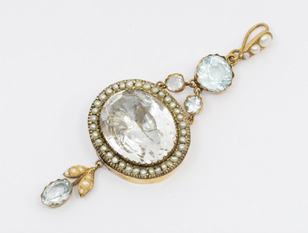 A Victorian gold pendant featuring a large oval rock crystal bordered by split seed pearls, flanked by pale blue aquamarine stones and leaf motifs. View IV
