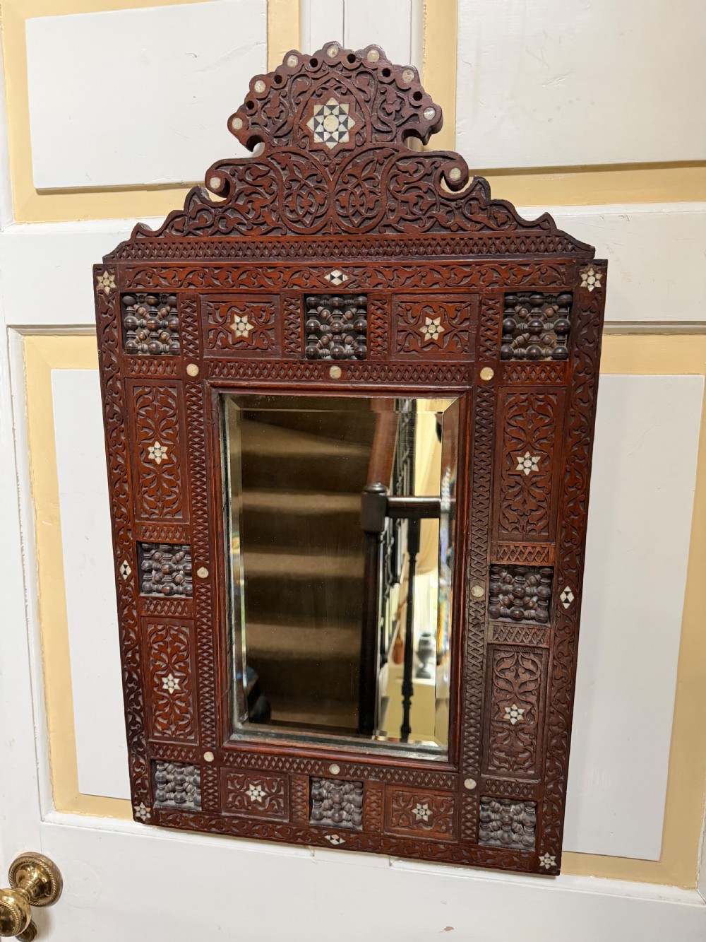 A late nineteenth-century carved dark wood wall mirror featuring geometric mother-of-pearl inlay, mashrabiya turned panels, and a shaped cresting. View II