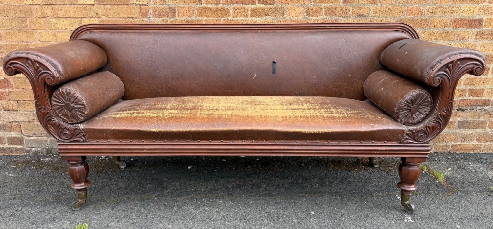 An early nineteenth-century William IV mahogany framed settee with worn brown leather upholstery, carved scroll arms, turned legs on brass castors, and matching bolster cushions.