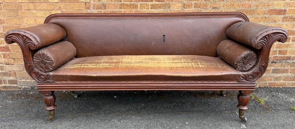 An early nineteenth-century William IV mahogany framed settee with worn brown leather upholstery, carved scroll arms, turned legs on brass castors, and matching bolster cushions. View II