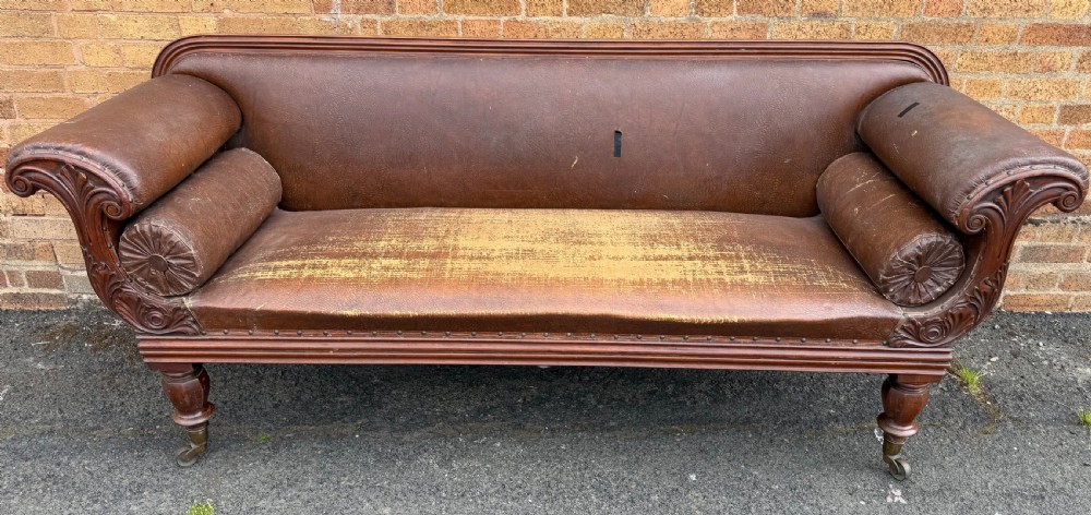 An early nineteenth-century William IV mahogany framed settee with worn brown leather upholstery, carved scroll arms, turned legs on brass castors, and matching bolster cushions. View III