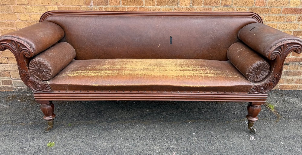 An early nineteenth-century William IV mahogany framed settee with worn brown leather upholstery, carved scroll arms, turned legs on brass castors, and matching bolster cushions. View IV