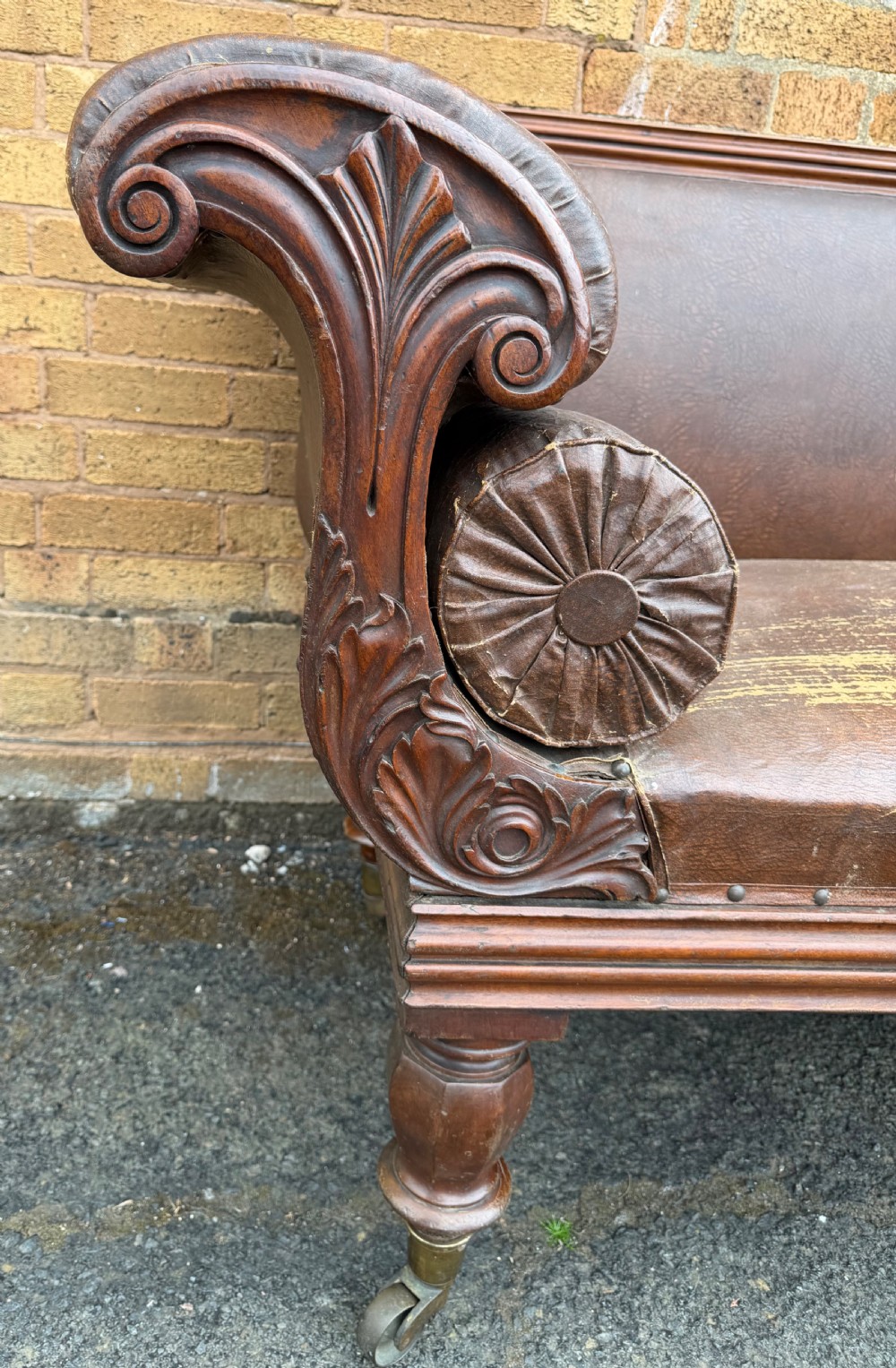 An early nineteenth-century William IV mahogany framed settee with worn brown leather upholstery, carved scroll arms, turned legs on brass castors, and matching bolster cushions. View V