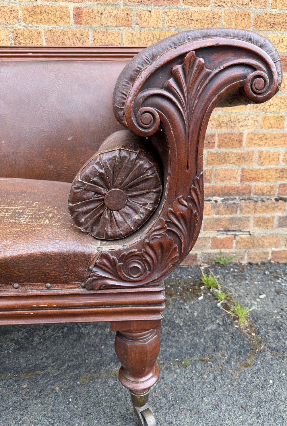 An early nineteenth-century William IV mahogany framed settee with worn brown leather upholstery, carved scroll arms, turned legs on brass castors, and matching bolster cushions. View X