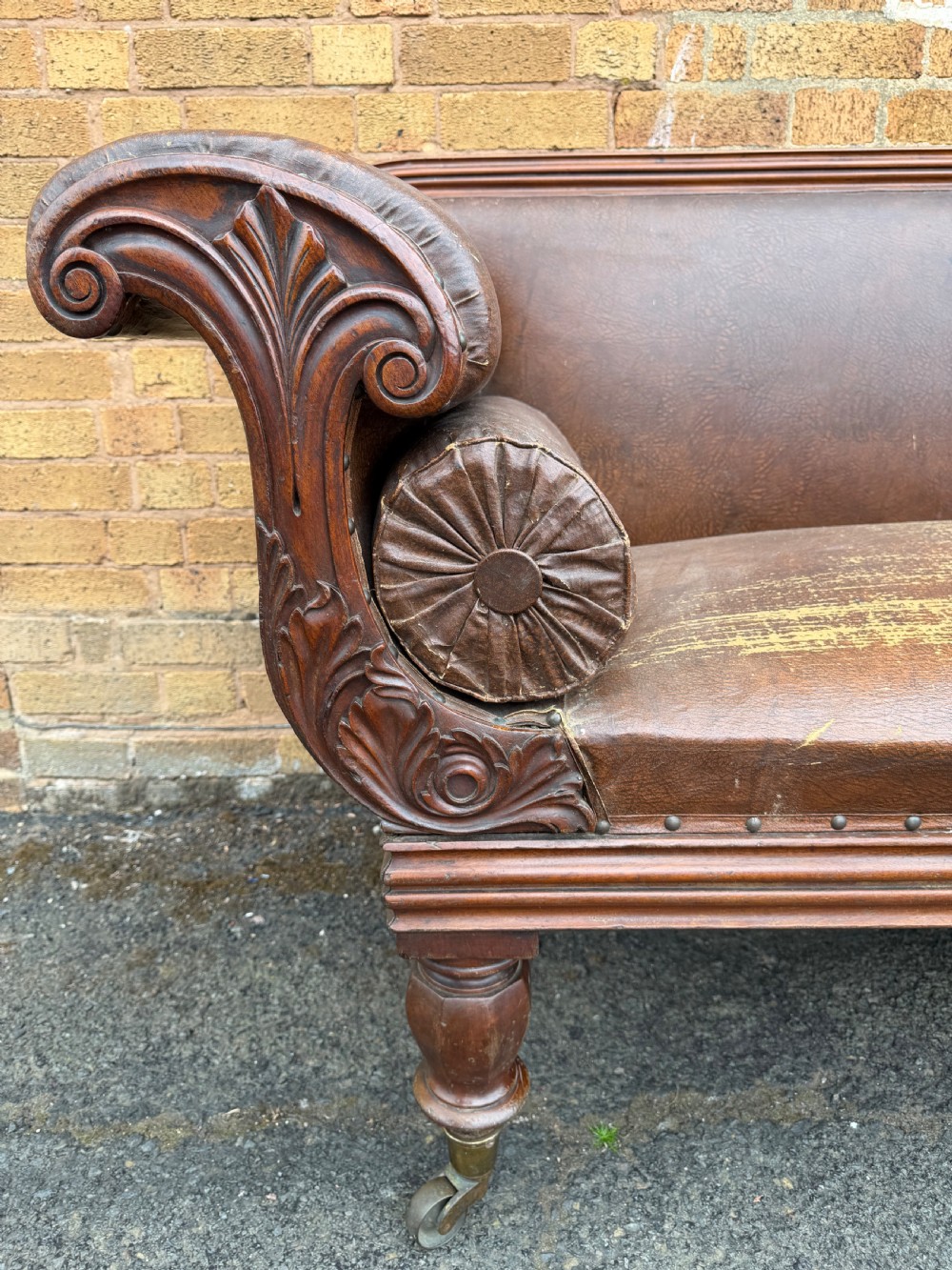 An early nineteenth-century William IV mahogany framed settee with worn brown leather upholstery, carved scroll arms, turned legs on brass castors, and matching bolster cushions. View XIV