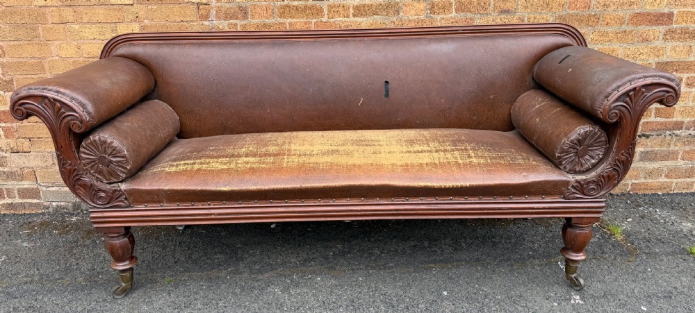 An early nineteenth-century William IV mahogany framed settee with worn brown leather upholstery, carved scroll arms, turned legs on brass castors, and matching bolster cushions. View XV