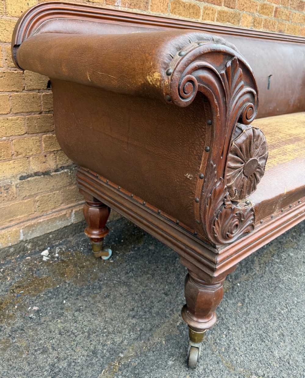 An early nineteenth-century William IV mahogany framed settee with worn brown leather upholstery, carved scroll arms, turned legs on brass castors, and matching bolster cushions. View XVI