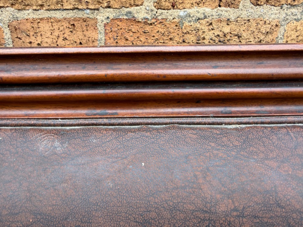 An early nineteenth-century William IV mahogany framed settee with worn brown leather upholstery, carved scroll arms, turned legs on brass castors, and matching bolster cushions. View XVIII