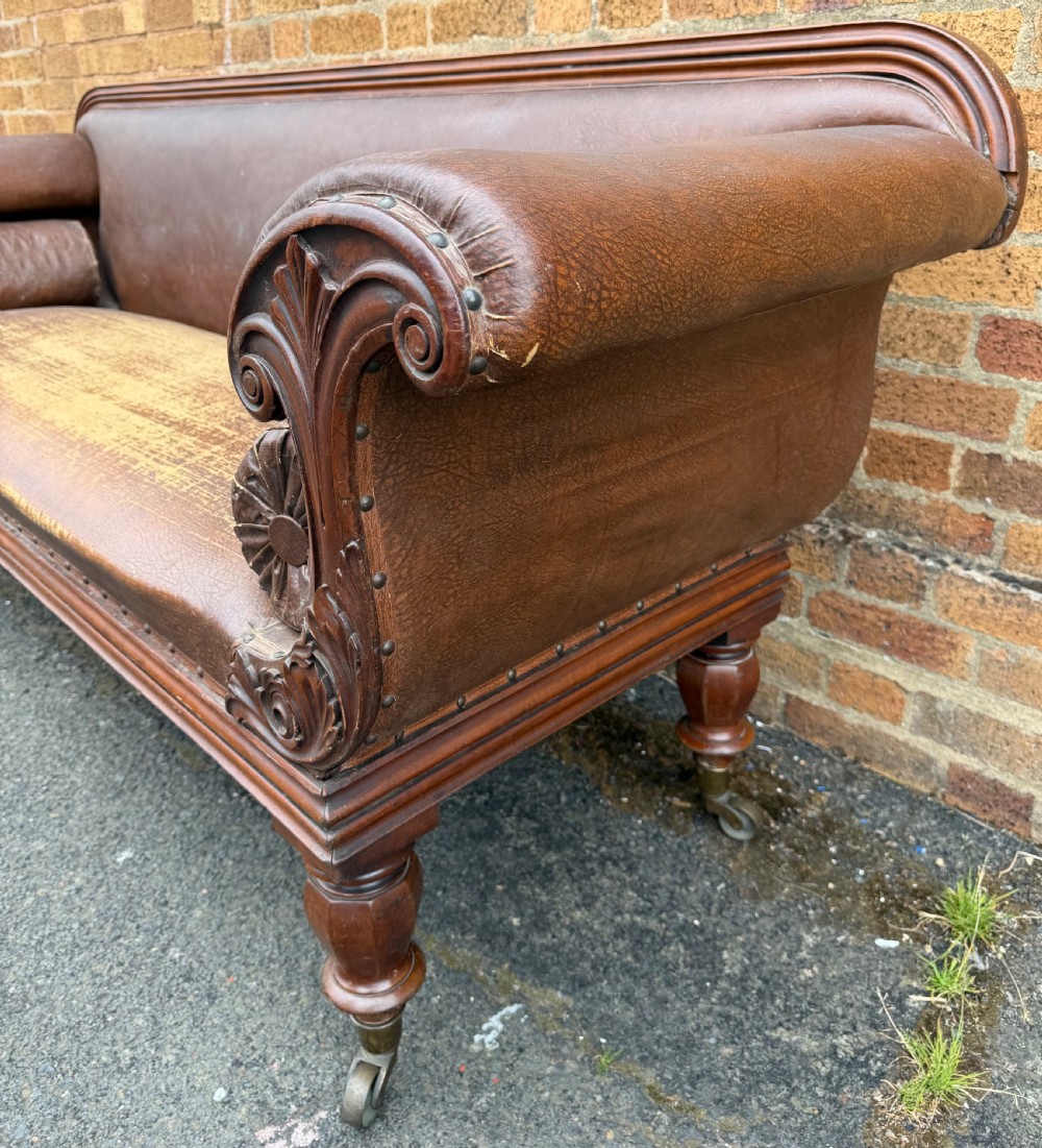 An early nineteenth-century William IV mahogany framed settee with worn brown leather upholstery, carved scroll arms, turned legs on brass castors, and matching bolster cushions. View XIX