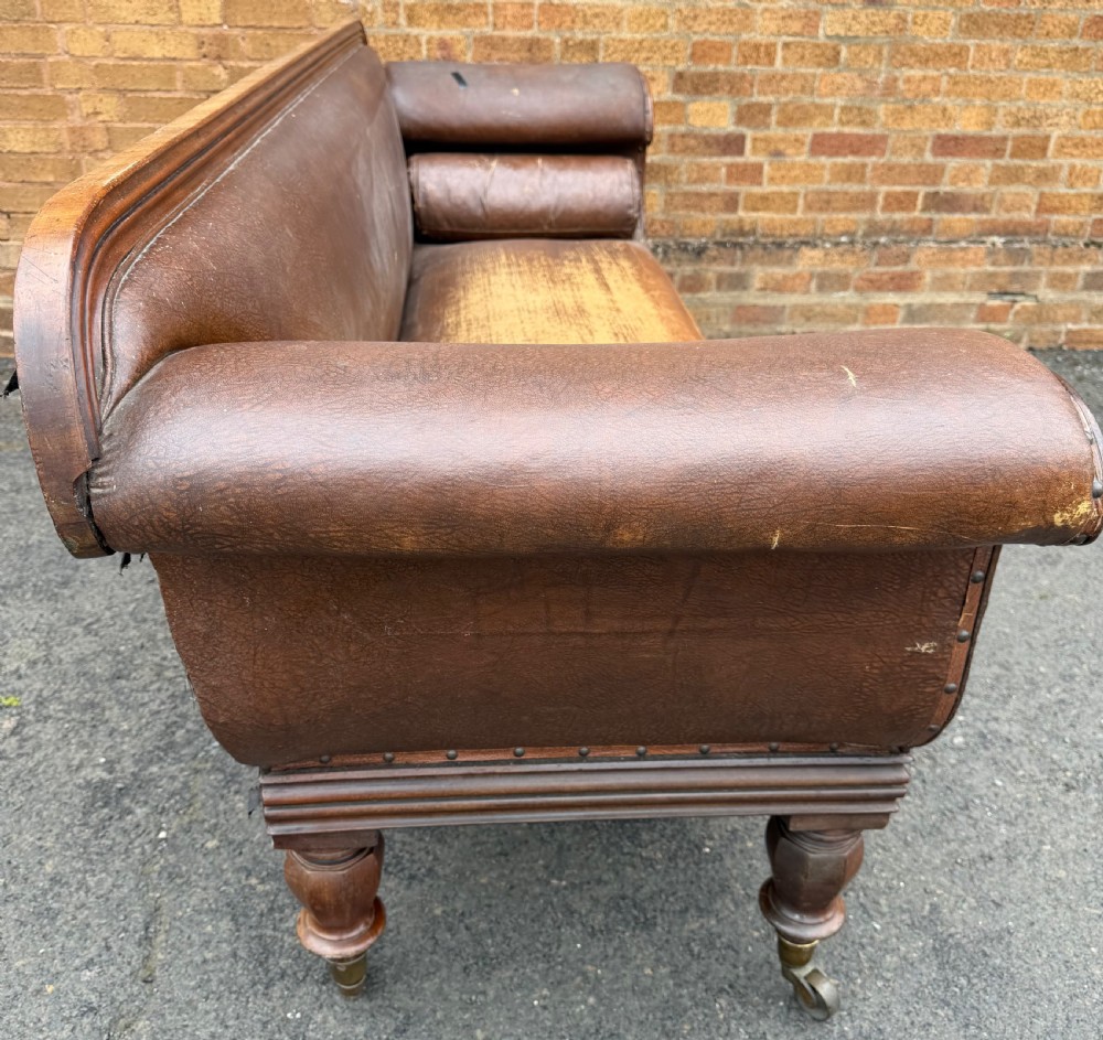 An early nineteenth-century William IV mahogany framed settee with worn brown leather upholstery, carved scroll arms, turned legs on brass castors, and matching bolster cushions. View XX