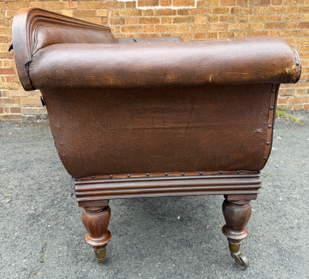 An early nineteenth-century William IV mahogany framed settee with worn brown leather upholstery, carved scroll arms, turned legs on brass castors, and matching bolster cushions. View XXI