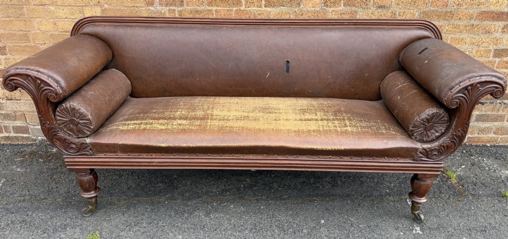 An early nineteenth-century William IV mahogany framed settee with worn brown leather upholstery, carved scroll arms, turned legs on brass castors, and matching bolster cushions. View XXIV