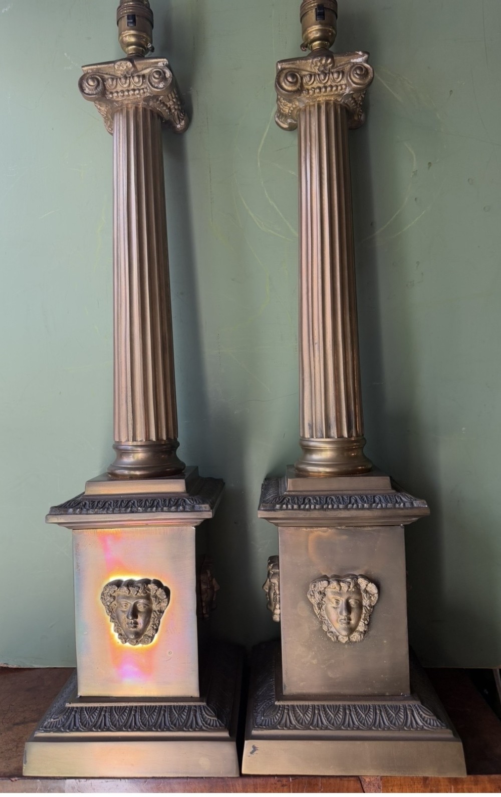 A pair of early twentieth-century brass table lamps featuring fluted classical columns, Corinthian capitals, and square pedestal bases decorated with cast face masks.